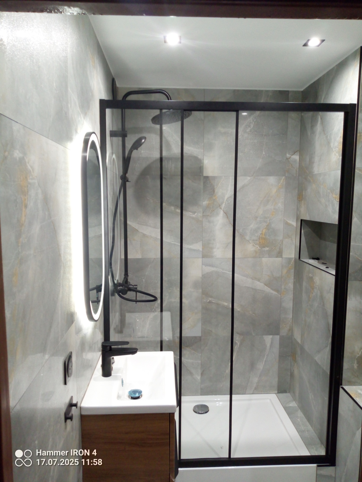 Nowoczesna łazienka z kabiną prysznicową typu walk-in, szare płytki imitujące marmur, podświetlane lustro i czarna armatura. Widoczne detale wykończenia.