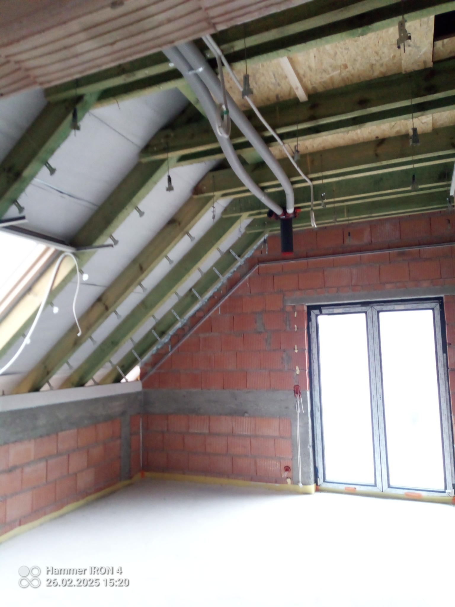 Poddasze w trakcie prac: widoczne drewniane krokwie, izolacja, instalacje wentylacyjne i okno. Ściany z czerwonej cegły. Etap przed wykończeniem.