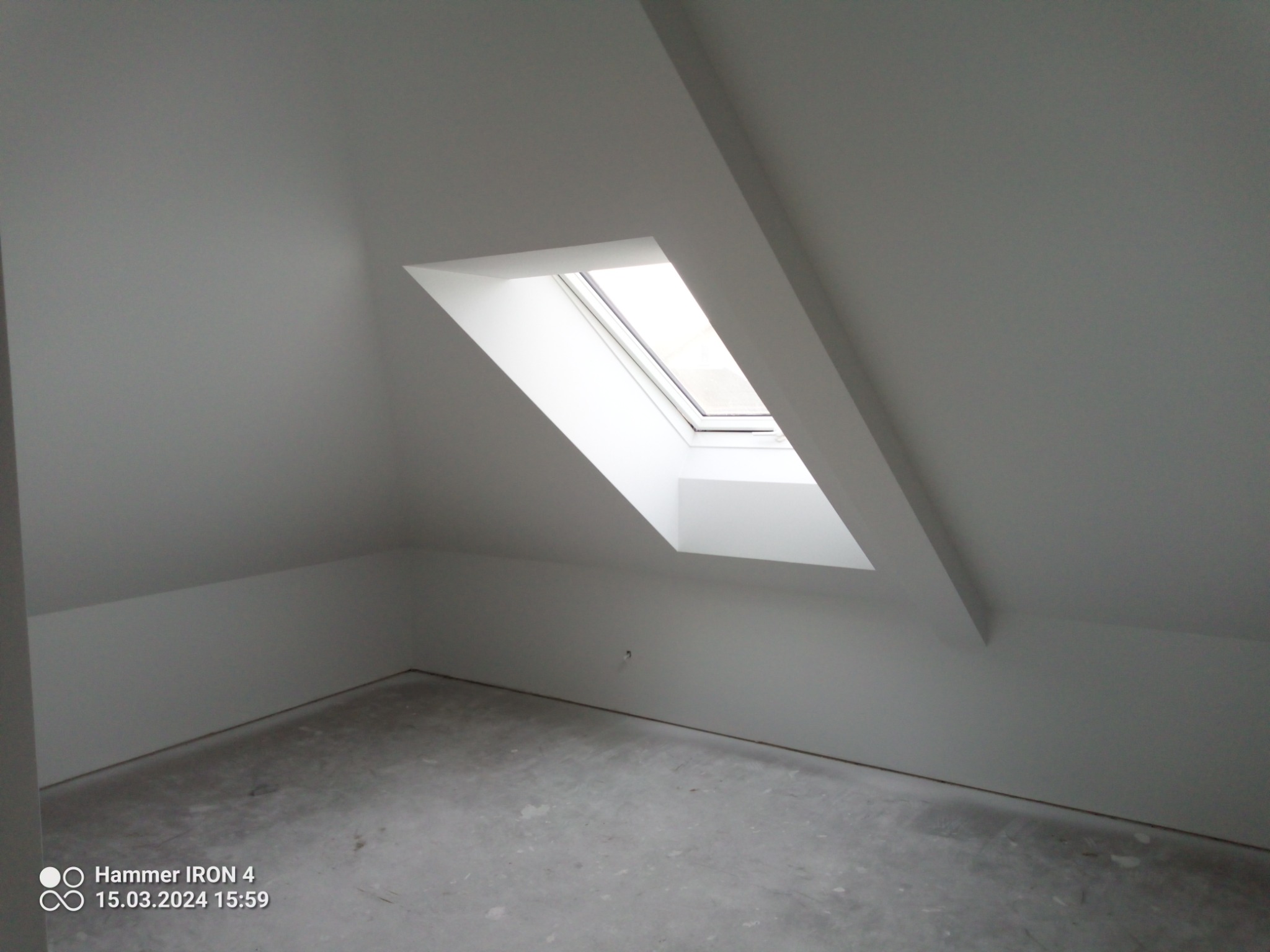 Poddasze po szpachlowaniu i malowaniu na biało, widoczne okno dachowe w skośnej ścianie, przygotowane do dalszych prac wykończeniowych.