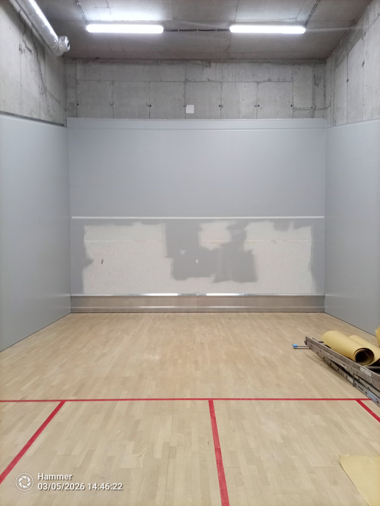 Wnętrze kortu do squasha w trakcie remontu: szpachlowana ściana, widoczne narzędzia i drabina. Podłoga z czerwonymi liniami. Surowy betonowy sufit z oświetleniem.