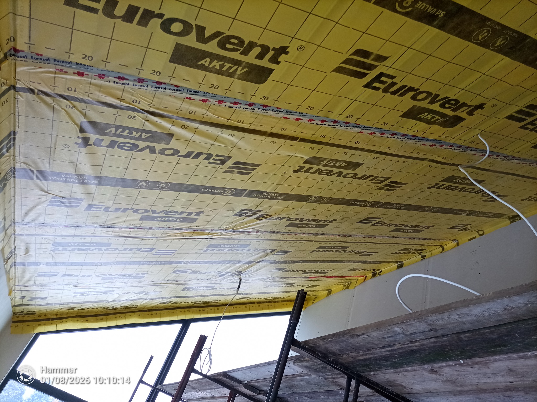 Poddasze z widoczną izolacją Eurovent Aktiv, zabezpieczoną taśmą EuraSol. Widoczne elementy konstrukcyjne i przewody. Ujęcie z dołu, z rusztowania.