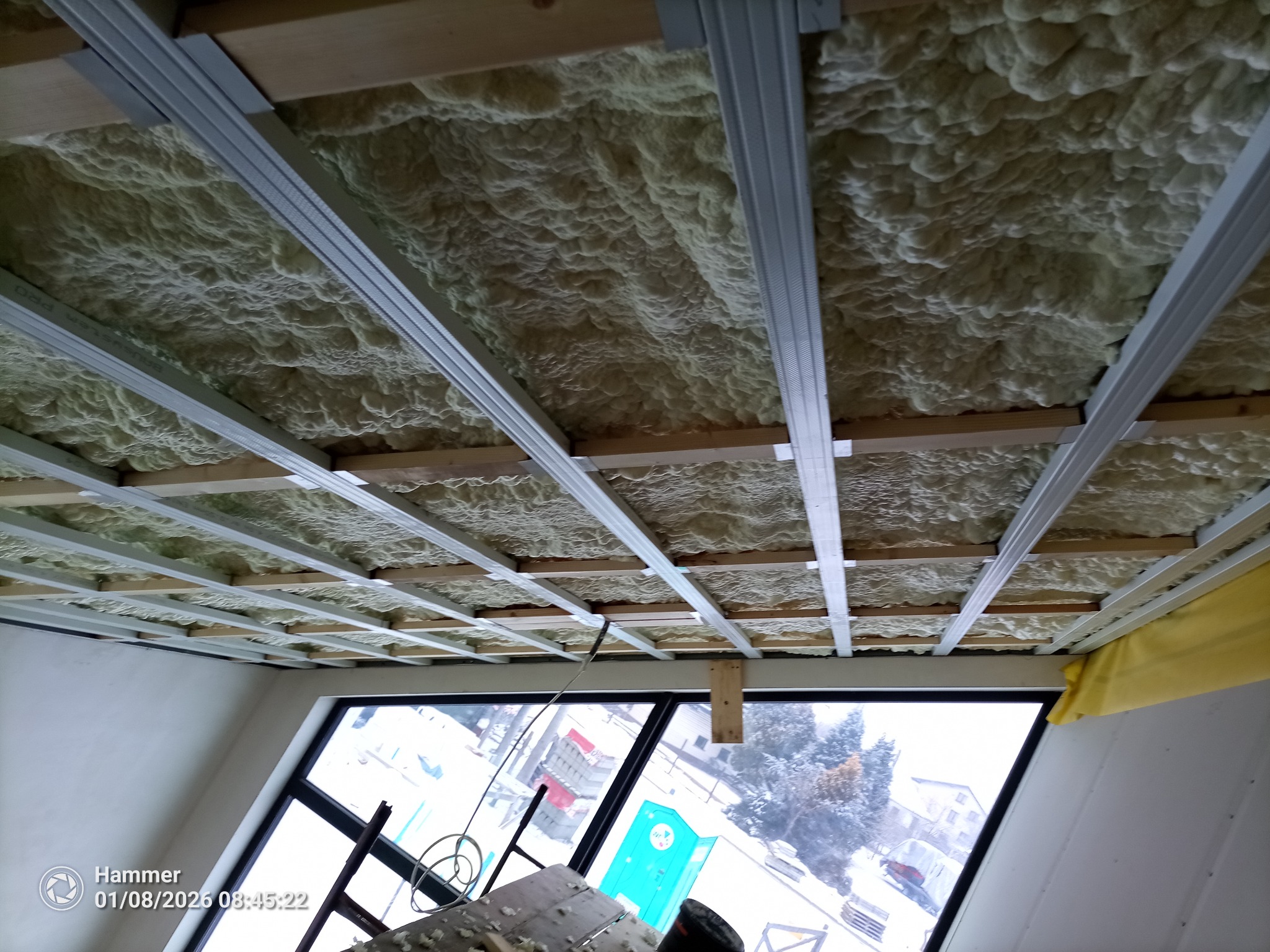 Poddasze z widoczną konstrukcją metalową i pianą izolacyjną, widok z dołu na okno. Etap ocieplania poddasza z użyciem piany PUR.