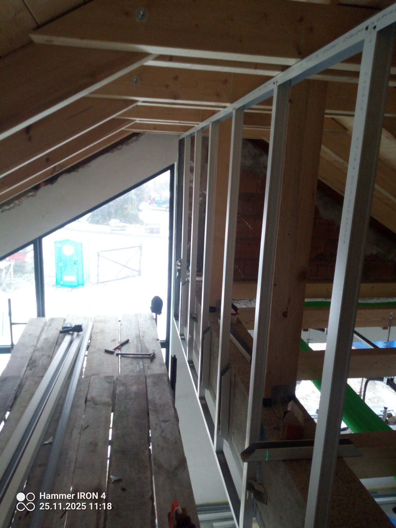 Montaż metalowej konstrukcji pod płyty G-K na poddaszu. Widoczne drewniane elementy więźby dachowej i narzędzia na prowizorycznym podeście. W tle okno z widokiem na zewnątrz.