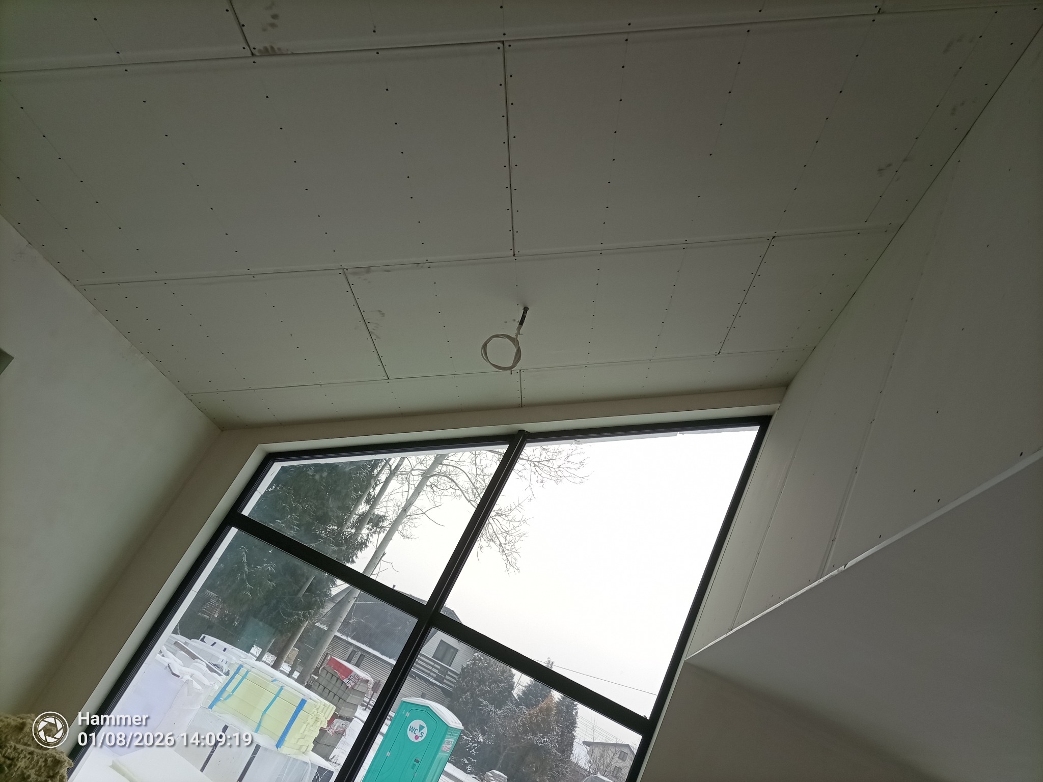 Sufit z płyt kartonowo-gipsowych w trakcie montażu, widoczne łączenia i wkręty. W tle okno z widokiem na zimowy krajobraz i materiały budowlane.