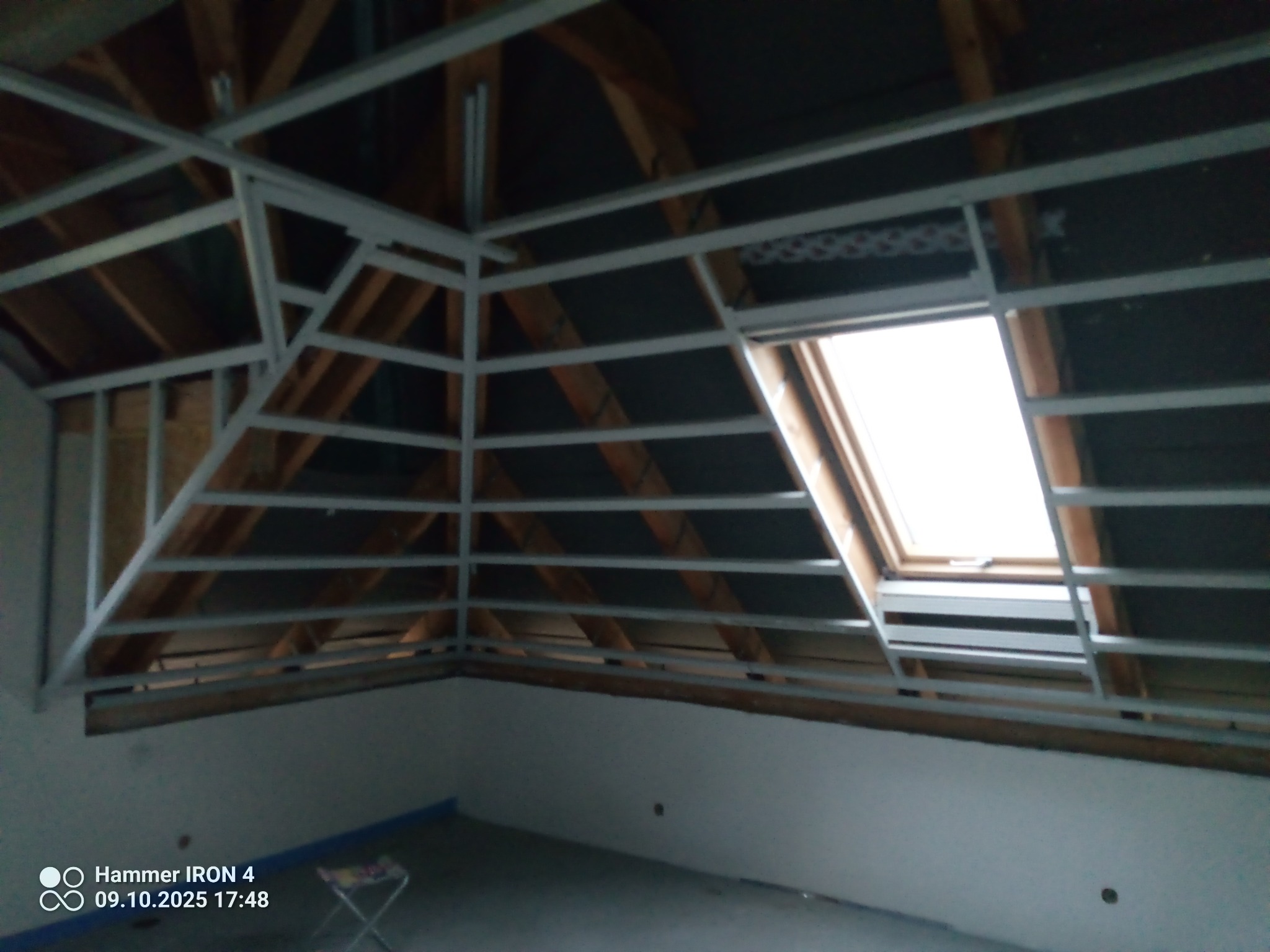 Szkielet metalowy pod zabudowę poddasza z oknem dachowym, widoczne drewniane elementy konstrukcyjne dachu i folia izolacyjna. Przygotowanie do montażu płyt.