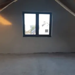 Poddasze po nałożeniu gładzi gipsowej; widoczne drewniane elementy konstrukcji dachu, okno z widokiem na budynek z kominem, świeżo wykończona ściana z delikatnym cieniem od słońca, surowa podłoga.