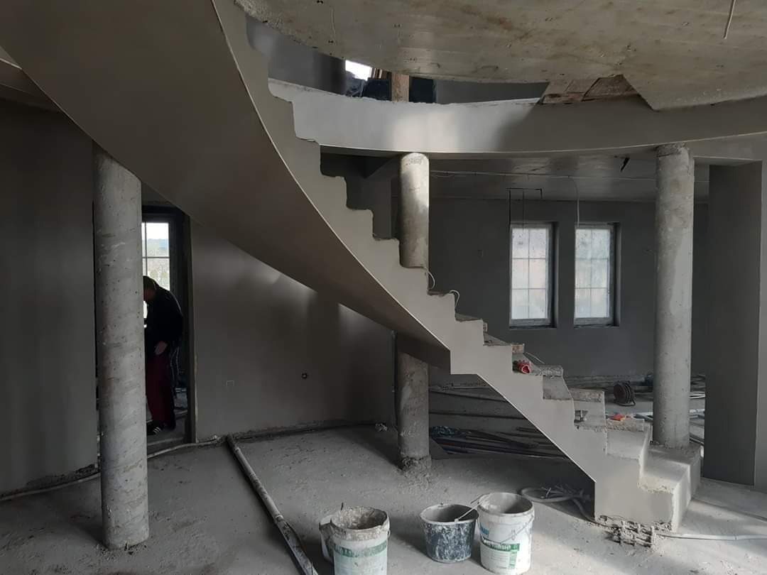 Wnętrze w trakcie tynkowania maszynowego: spiralne schody, kolumny i ściany pokryte świeżym tynkiem, widoczne wiadra i narzędzia, surowy stan wykończenia.