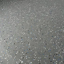 Szara, gładka powierzchnia podłogi z widocznymi, nieregularnymi płatkami w kolorach białym, czarnym i niebieskim, tworzącymi wzór przypominający terrazzo.