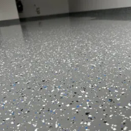 Szara posadzka żywiczna z drobnymi, nieregularnymi płatkami w kolorach białym, czarnym i niebieskim, tworzącymi wzór terrazzo, z widocznym połyskiem i odbiciami światła.