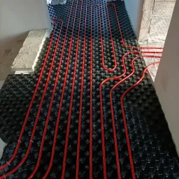 Czerwone rury ogrzewania podłogowego ułożone na czarnej macie systemowej w trakcie montażu, widoczne fragmenty ścian w surowym stanie.