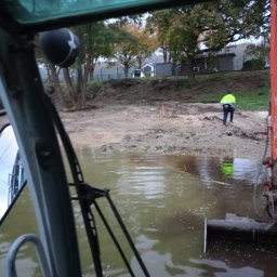 MARTRANS Michał Marciński - Widok z kabiny maszyny budowlanej na teren z błotnistą wodą i pracownikiem w odblaskowej kamizelce na brzegu; w tle drzewa i budynki.