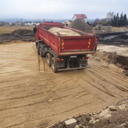 MARTRANS Michał Marciński - Czerwona ciężarówka Mercedes-Benz z piaskiem na placu budowy, widoczne ślady opon na piasku i pryzma kruszywa w tle.