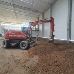 MARTRANS Michał Marciński - Czerwona koparka Atlas 1305 M w hali w Rumi wyrównuje teren pod posadzkę, w tle widoczny pracownik z niwelatorem.