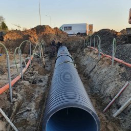 MARTRANS Michał Marciński - Instalacja dużej, karbowanej rury kanalizacyjnej w wykopie, widoczni dwaj pracownicy, pomarańczowe rury i szpadle w tle, firmowy van zaparkowany w oddali.