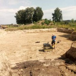 MARTRANS Michał Marciński - Pracownik obsługuje zagęszczarkę stopową na piaszczystym terenie wykopu pod fundamenty, z widocznymi śladami po maszynie i nierównościami podłoża, w tle drzewa i niebo.