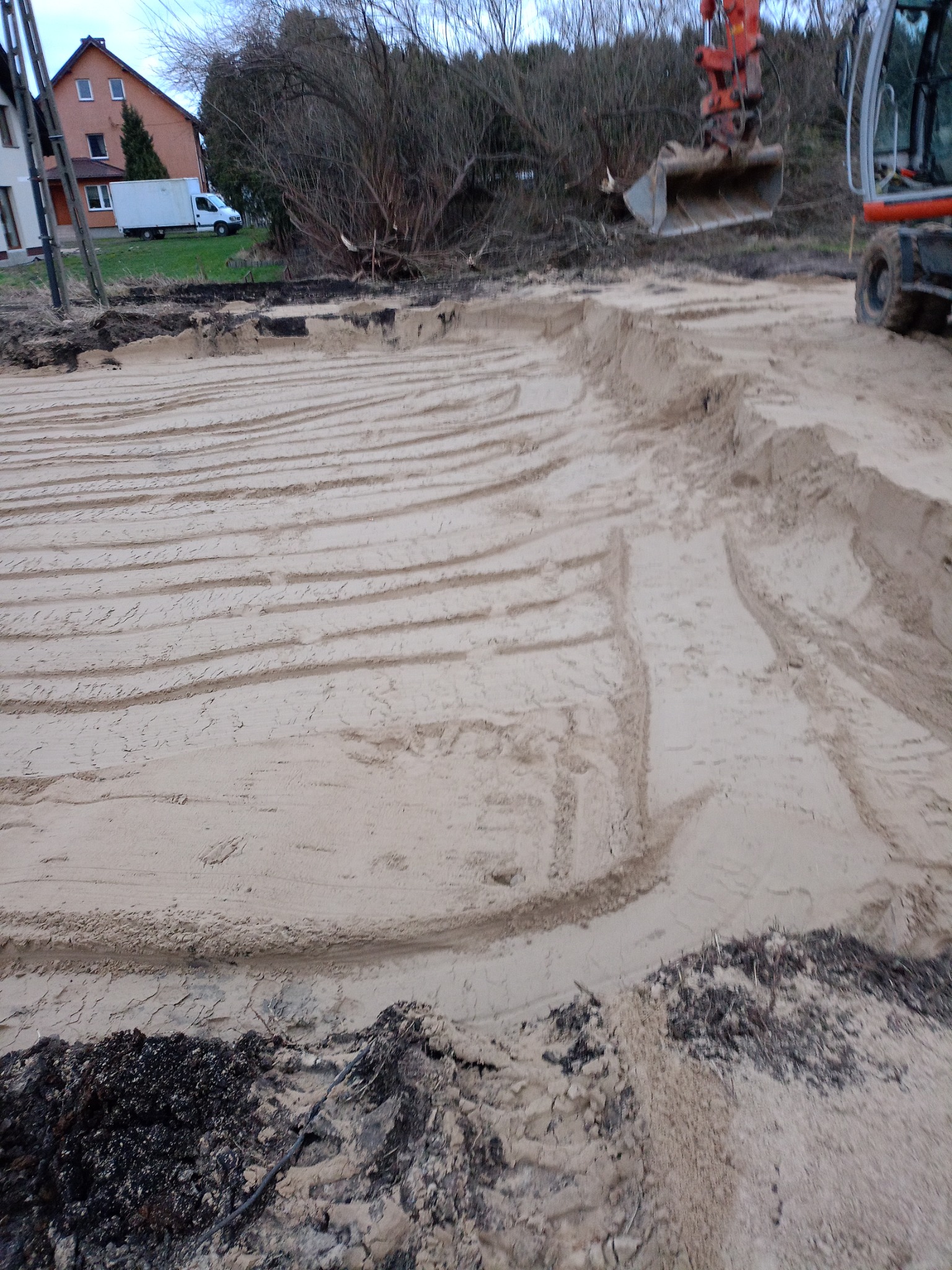 Nierówny teren z wyżłobieniami po maszynach budowlanych, w tle widoczny budynek i częściowo wykopany teren.