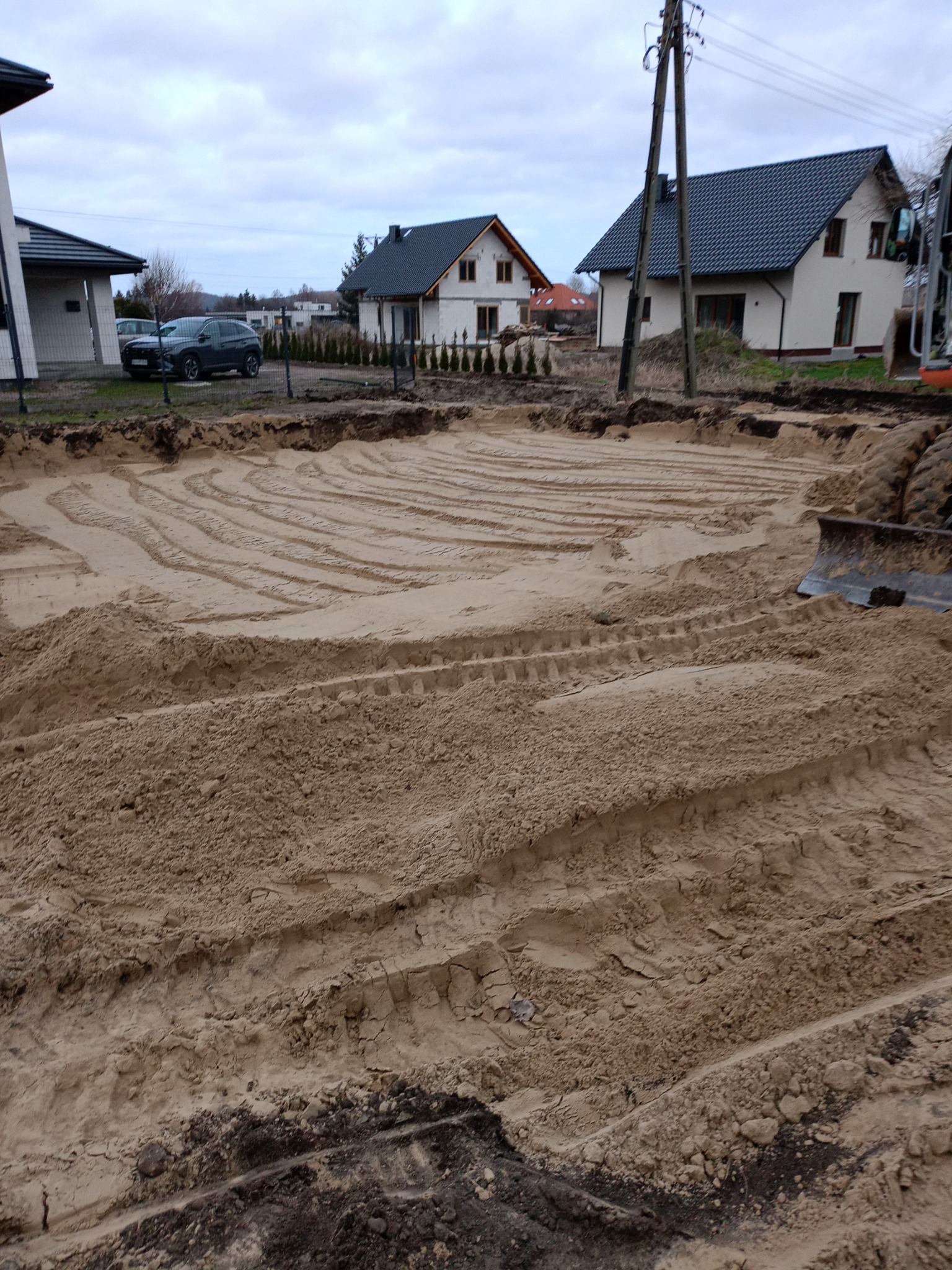 Przygotowany teren pod budowę z widocznymi śladami opon i wyrównanym piaskiem, w tle domy jednorodzinne i pochmurne niebo.