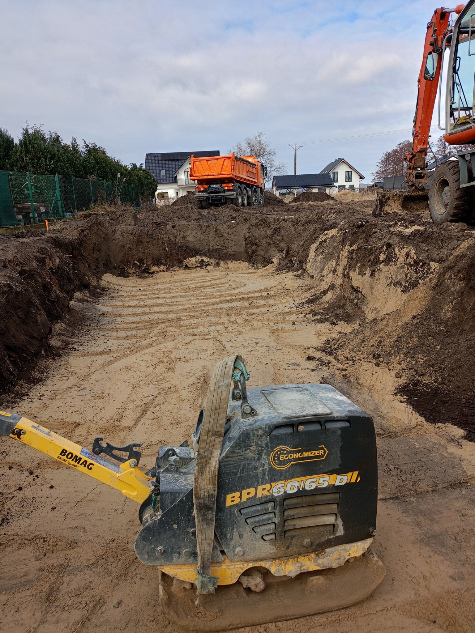 Pomarańczowa wywrotka stoi na skraju wykopu pod budowę, w tle domy jednorodzinne, na pierwszym planie ubijarka wibro płytowa BOMAG BPR60/65D.