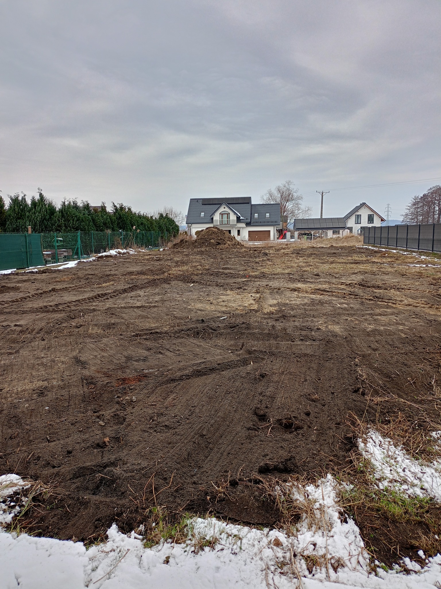 Przygotowany teren pod budowę domu, widoczne ślady opon i niewielki kopiec ziemi, w tle zabudowa jednorodzinna i pochmurne niebo.