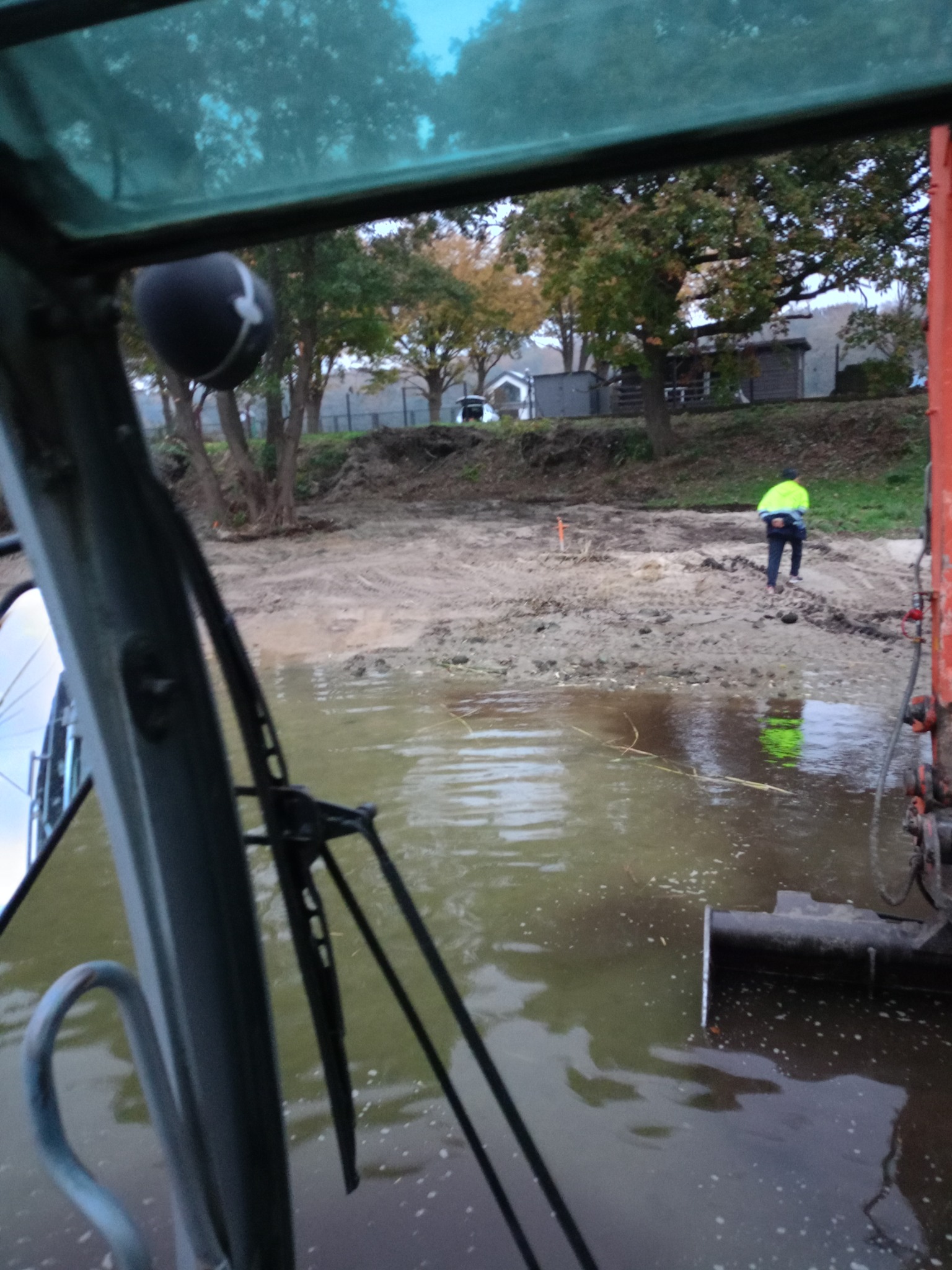 Widok z kabiny maszyny budowlanej na teren z błotnistą wodą i pracownikiem w odblaskowej kamizelce na brzegu; w tle drzewa i budynki.