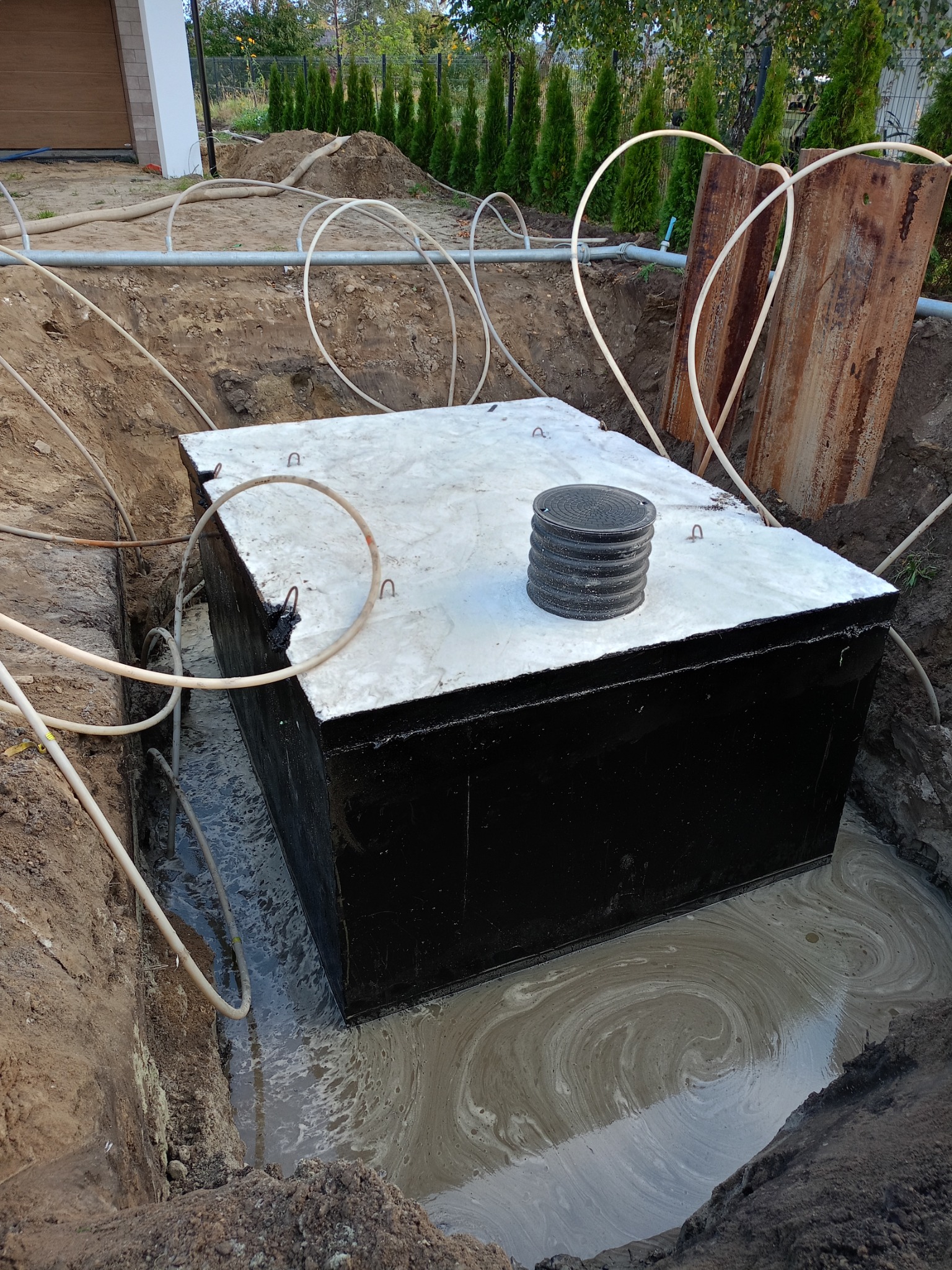 Czarny, betonowy zbiornik z pokrywą umieszczony w wykopie wypełnionym wodą, widoczne rury i metalowe szalunki, w tle zieleń.