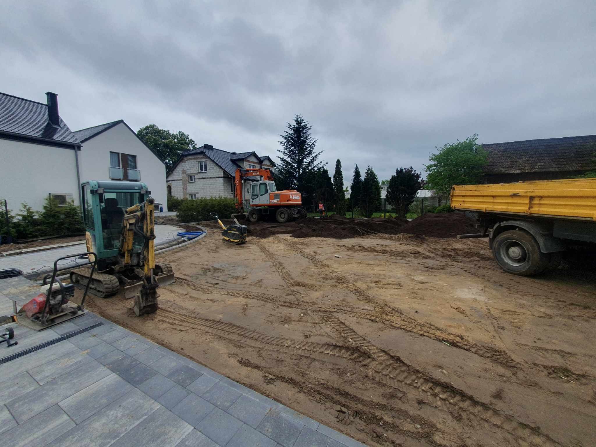 Przygotowanie terenu pod budowę: mini koparka, koparka Atlas, ubijak i wywrotka na placu budowy z częściowo ułożoną kostką brukową w tle.