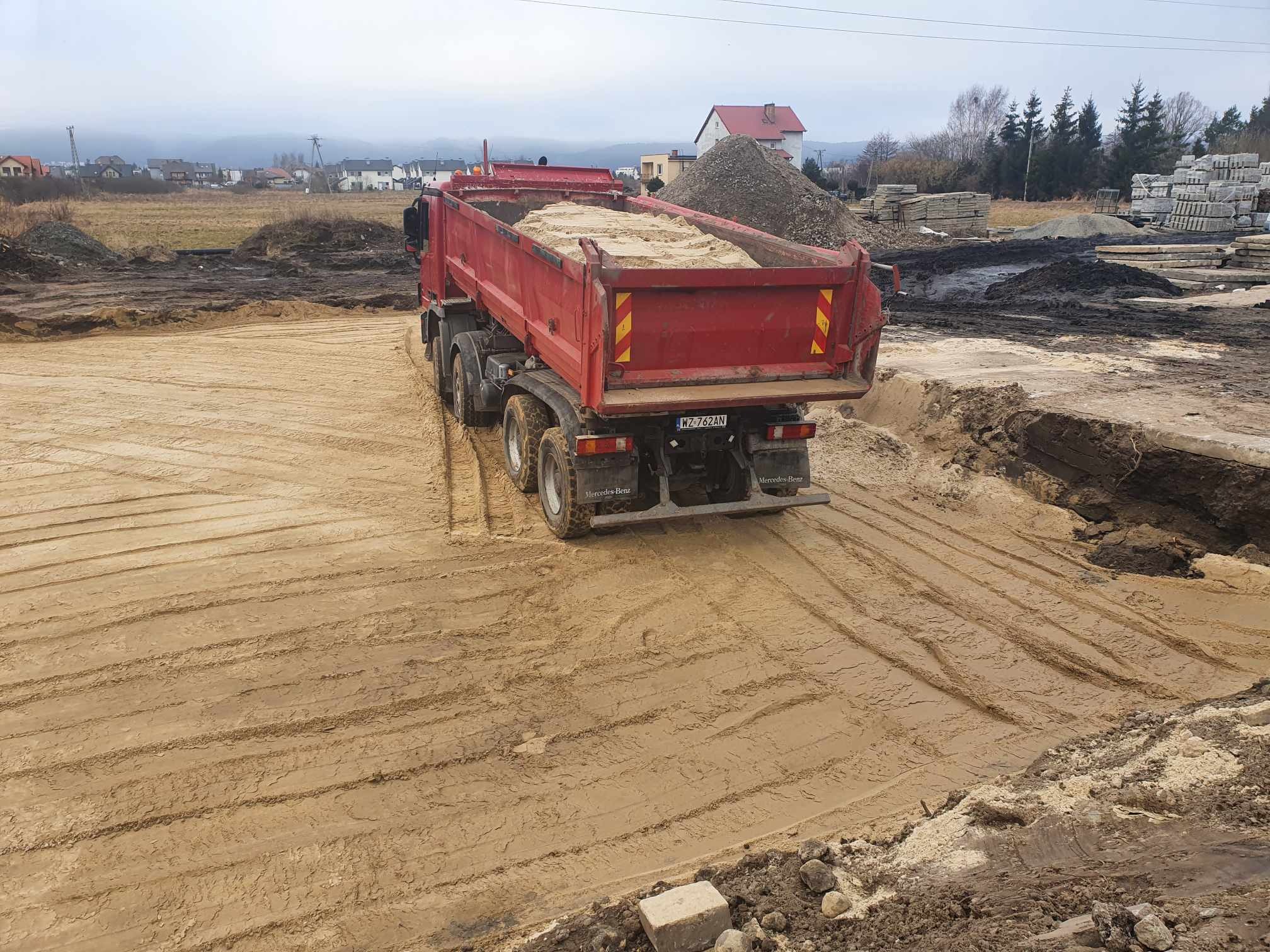 Czerwona ciężarówka Mercedes-Benz z piaskiem na placu budowy, widoczne ślady opon na piasku i pryzma kruszywa w tle.