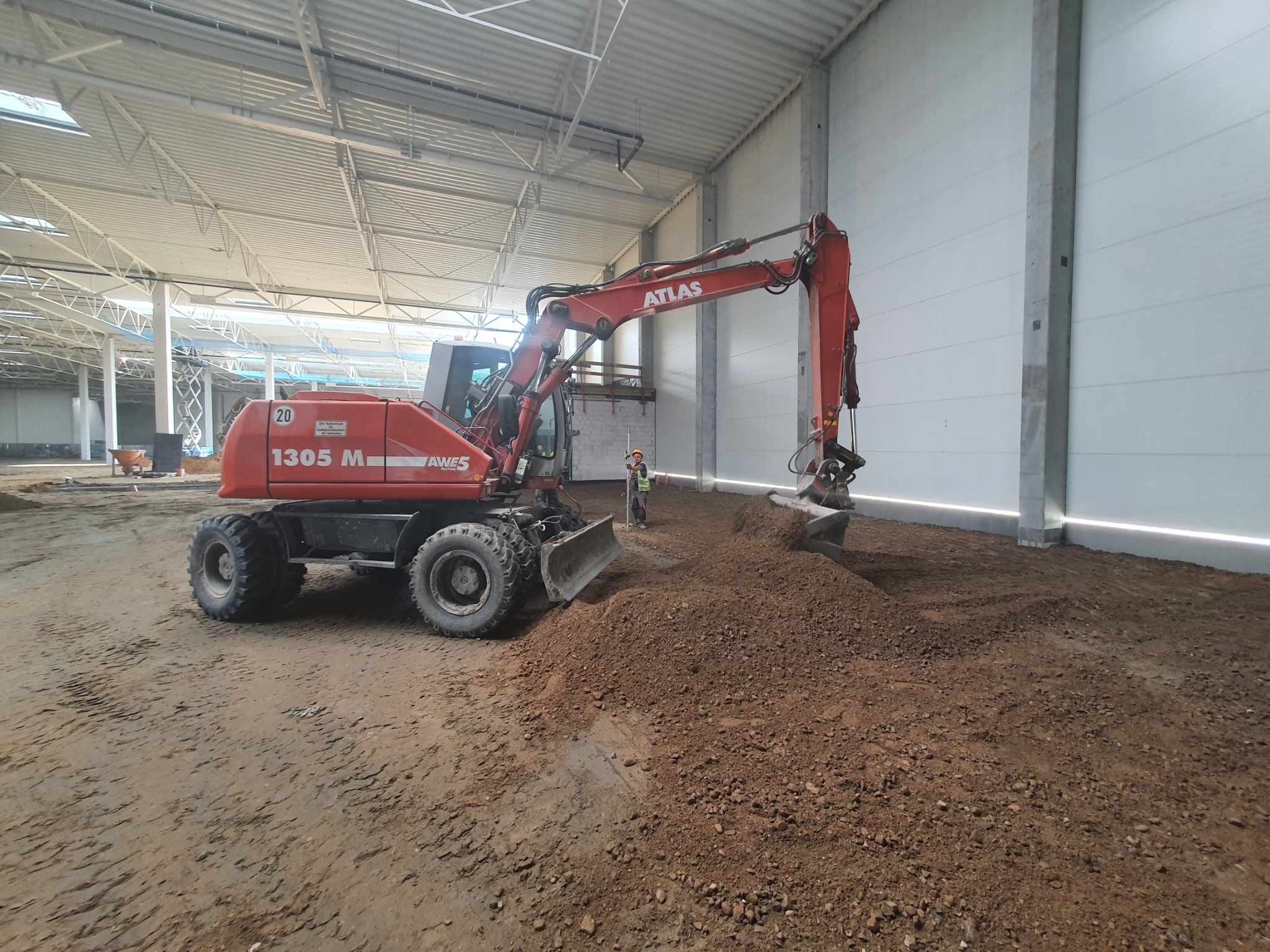 Czerwona koparka Atlas 1305 M w hali w Rumi wyrównuje teren pod posadzkę, w tle widoczny pracownik z niwelatorem.