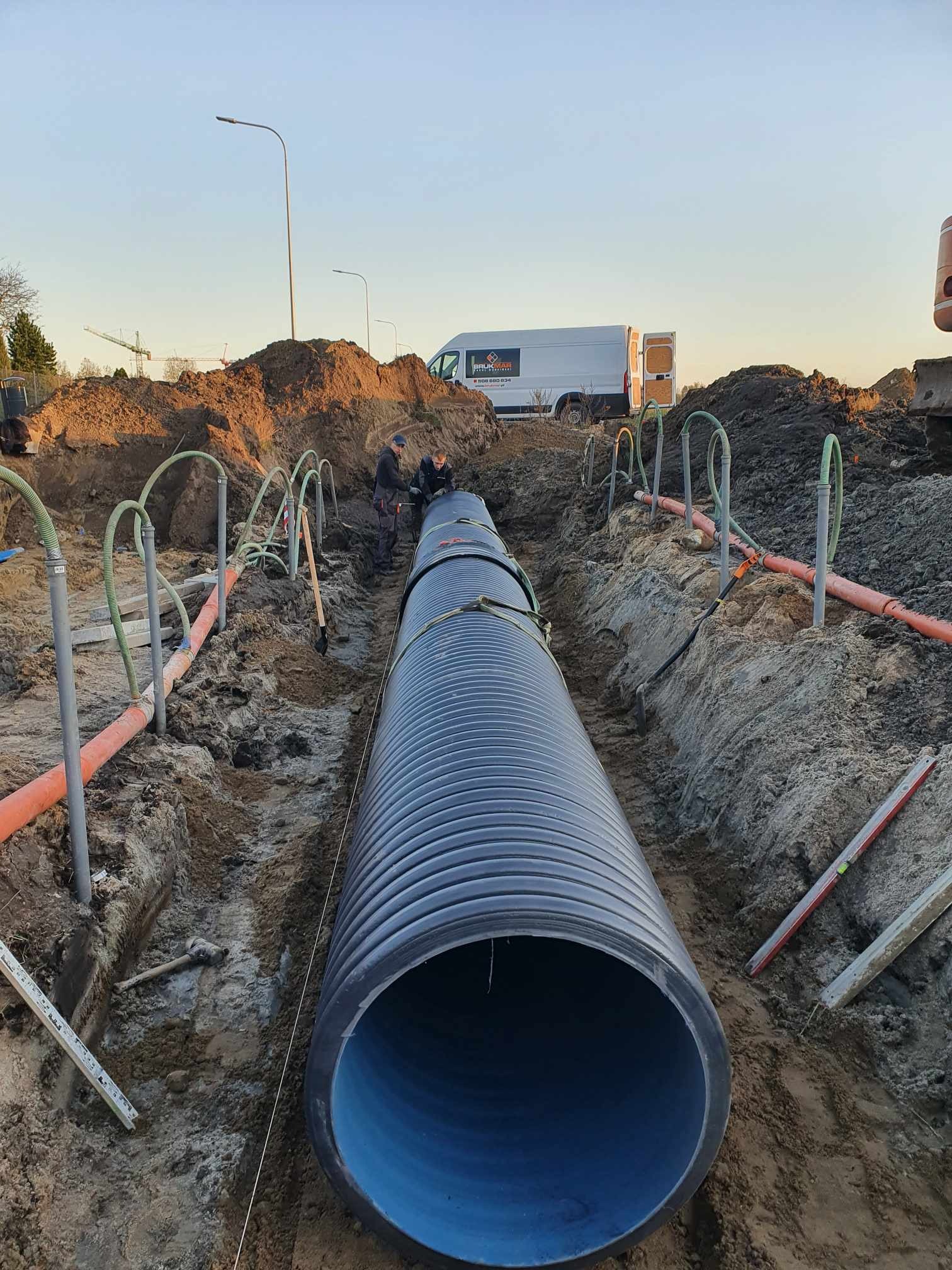 Instalacja dużej, karbowanej rury kanalizacyjnej w wykopie, widoczni dwaj pracownicy, pomarańczowe rury i szpadle w tle, firmowy van zaparkowany w oddali.