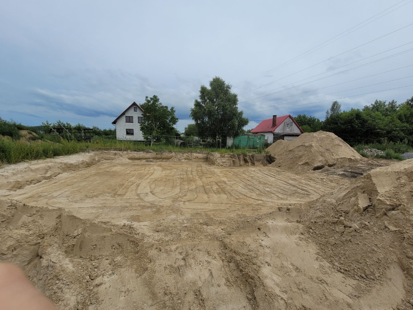 Przygotowany teren pod budowę domu jednorodzinnego, widoczne ślady po maszynach, usypane pryzmy ziemi, w tle dwa domy i linia wysokiego napięcia.