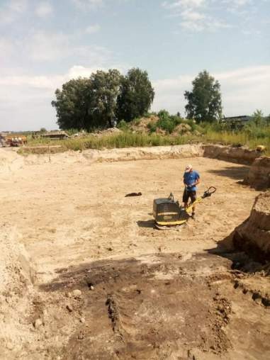 Pracownik obsługuje zagęszczarkę stopową na piaszczystym terenie wykopu pod fundamenty, z widocznymi śladami po maszynie i nierównościami podłoża, w tle drzewa i niebo.