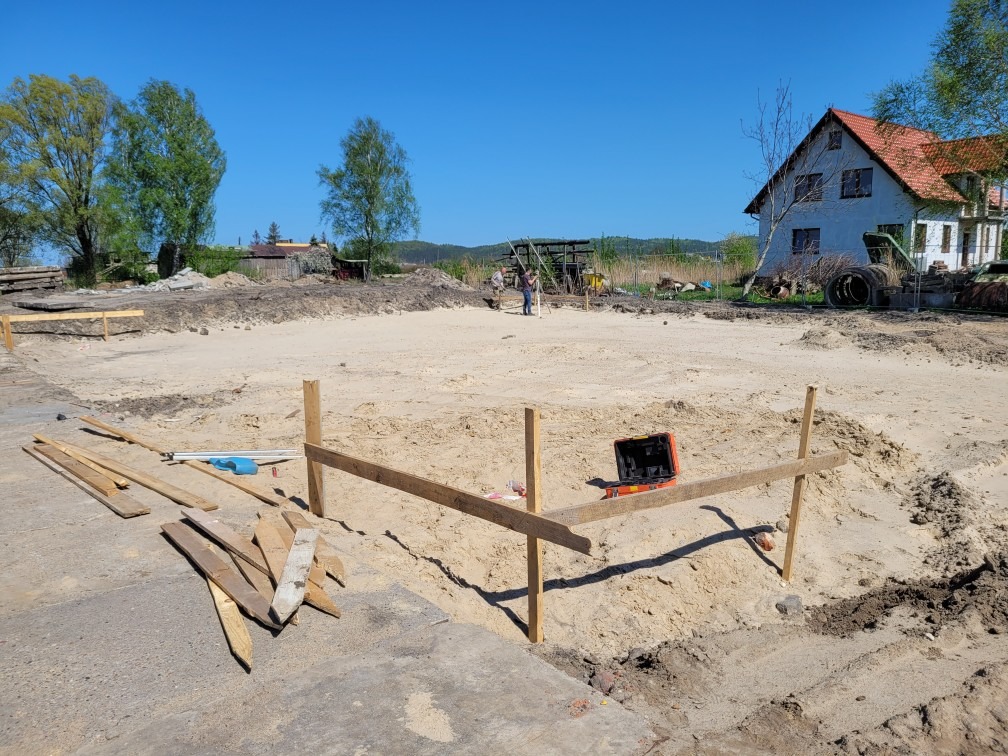 Przygotowany teren pod budowę z wytyczonymi fundamentami, widoczne drewniane słupki i poziomica, w tle budynek mieszkalny i drzewa.