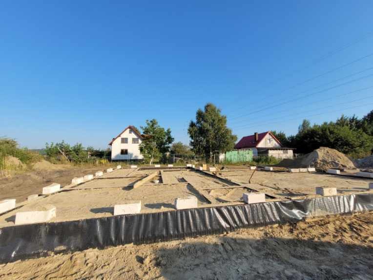 Przygotowany teren pod budowę domu jednorodzinnego, widoczne fundamenty zabezpieczone folią, w tle dwa domy i błękitne niebo.