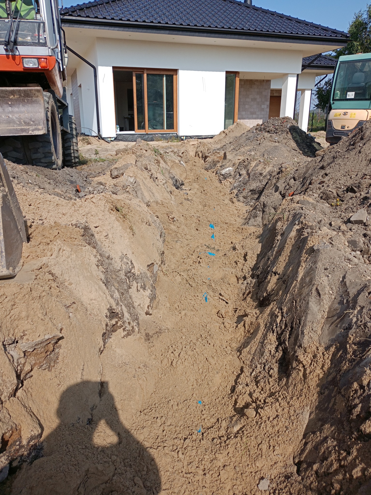 Długi, świeżo wykopany rów na piaszczystej glebie, z widocznymi fragmentami niebieskiej taśmy, obok domu jednorodzinnego w trakcie budowy i maszyn budowlanych.