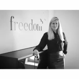 Portret kobiety w czarnej bluzce i spódnicy, stojącej w biurze z napisem 'freedom' na ścianie i kluczami w dłoni, czarno-białe zdjęcie.