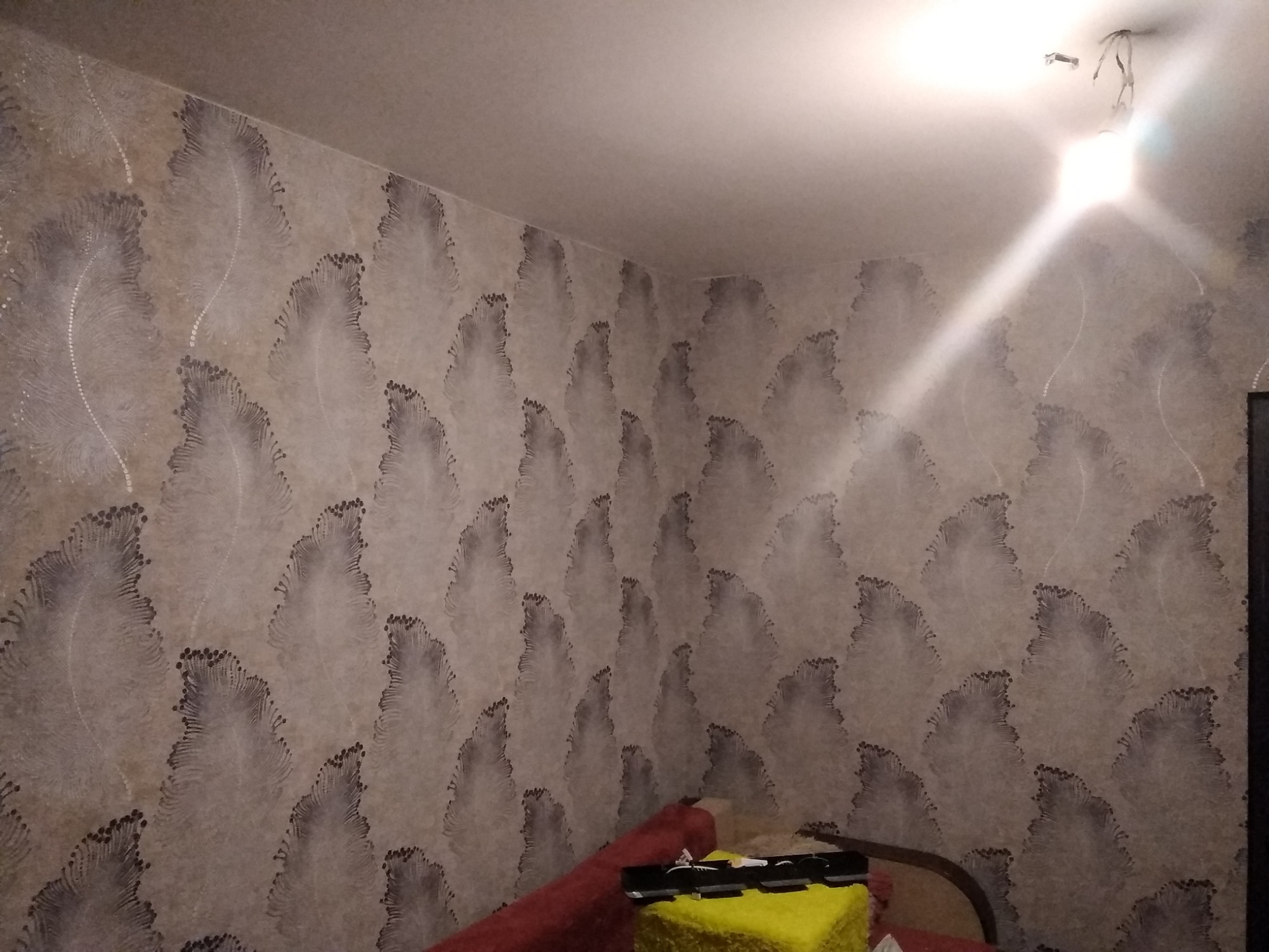 Ściana pokryta tapetą w motywie piór z niedokończonym oświetleniem sufitowym, widoczne fragmenty mebli i akcesoriów malarskich.
