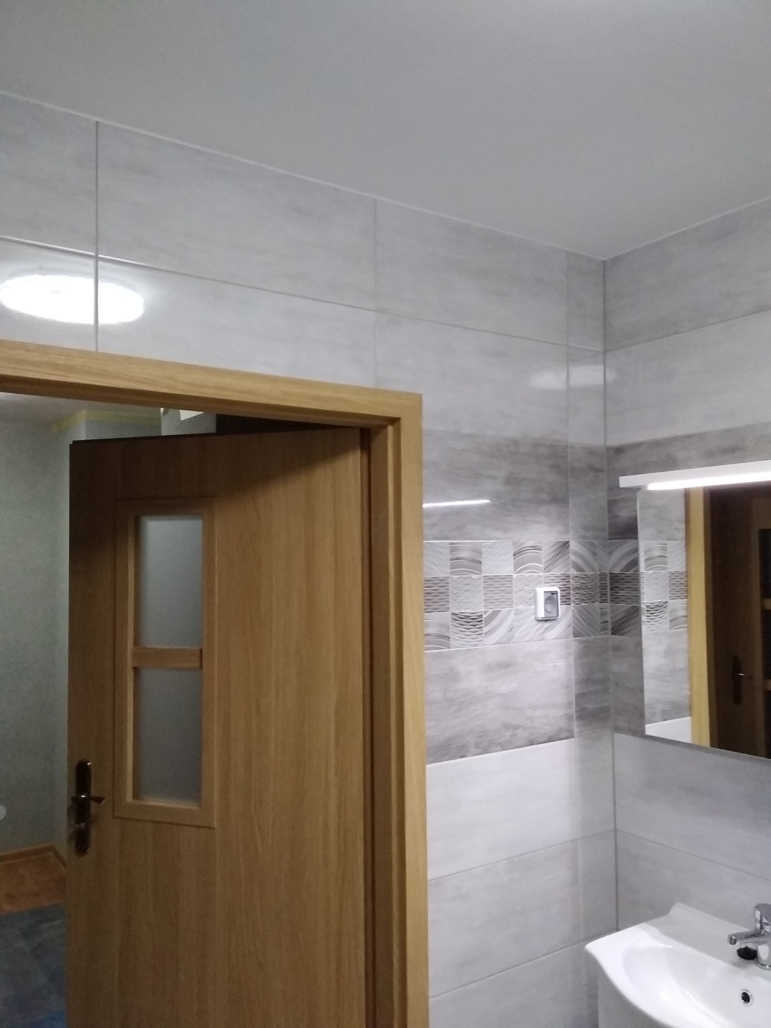 Wykończona łazienka z szaro-białymi płytkami, drewnianą ościeżnicą drzwiową i lustrem odbijającym fragment wnętrza