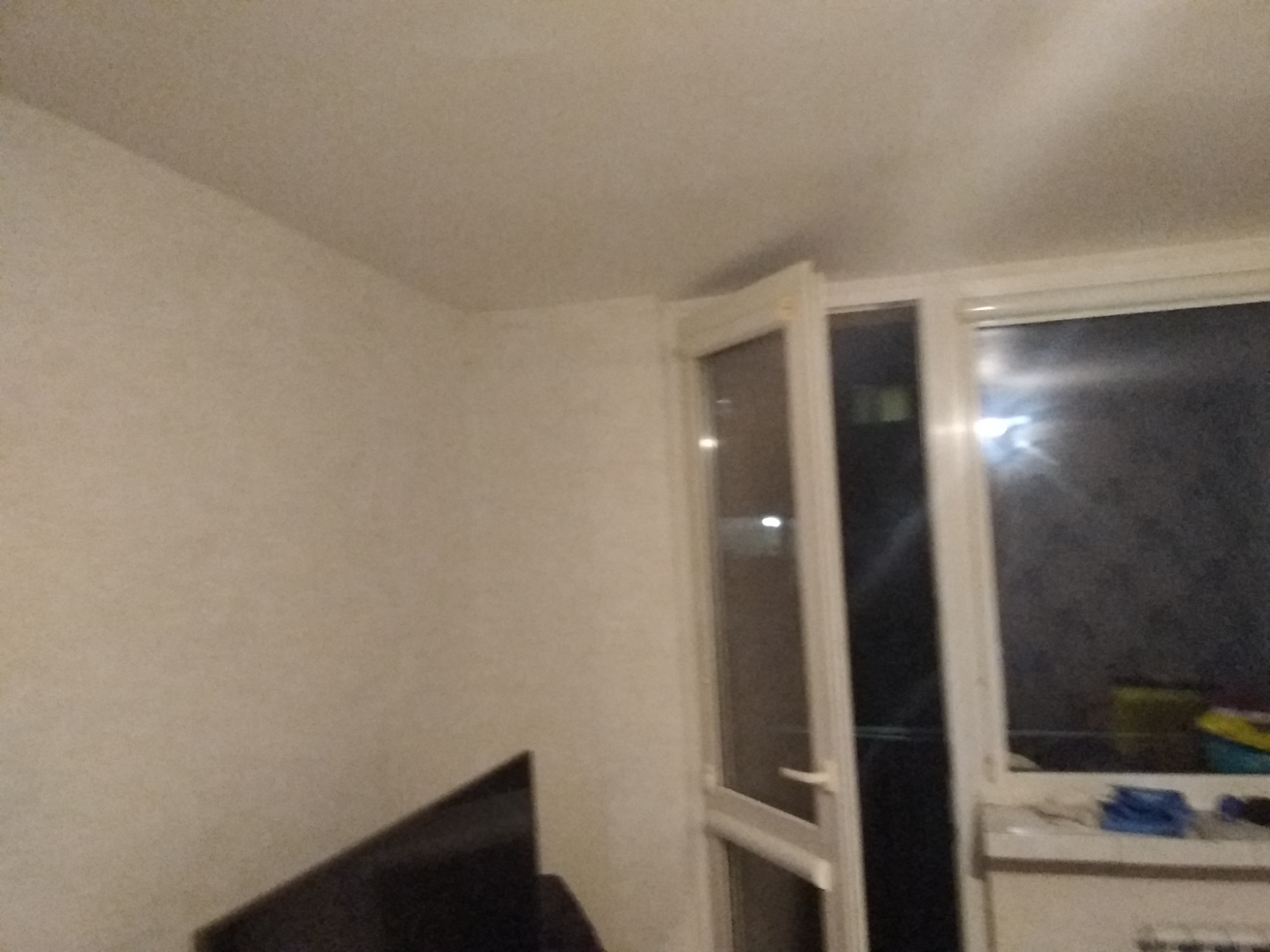 Wnętrze pokoju z otwartymi drzwiami balkonowymi, widoczna ściana z delikatną fakturą i fragment telewizora w rogu kadru.