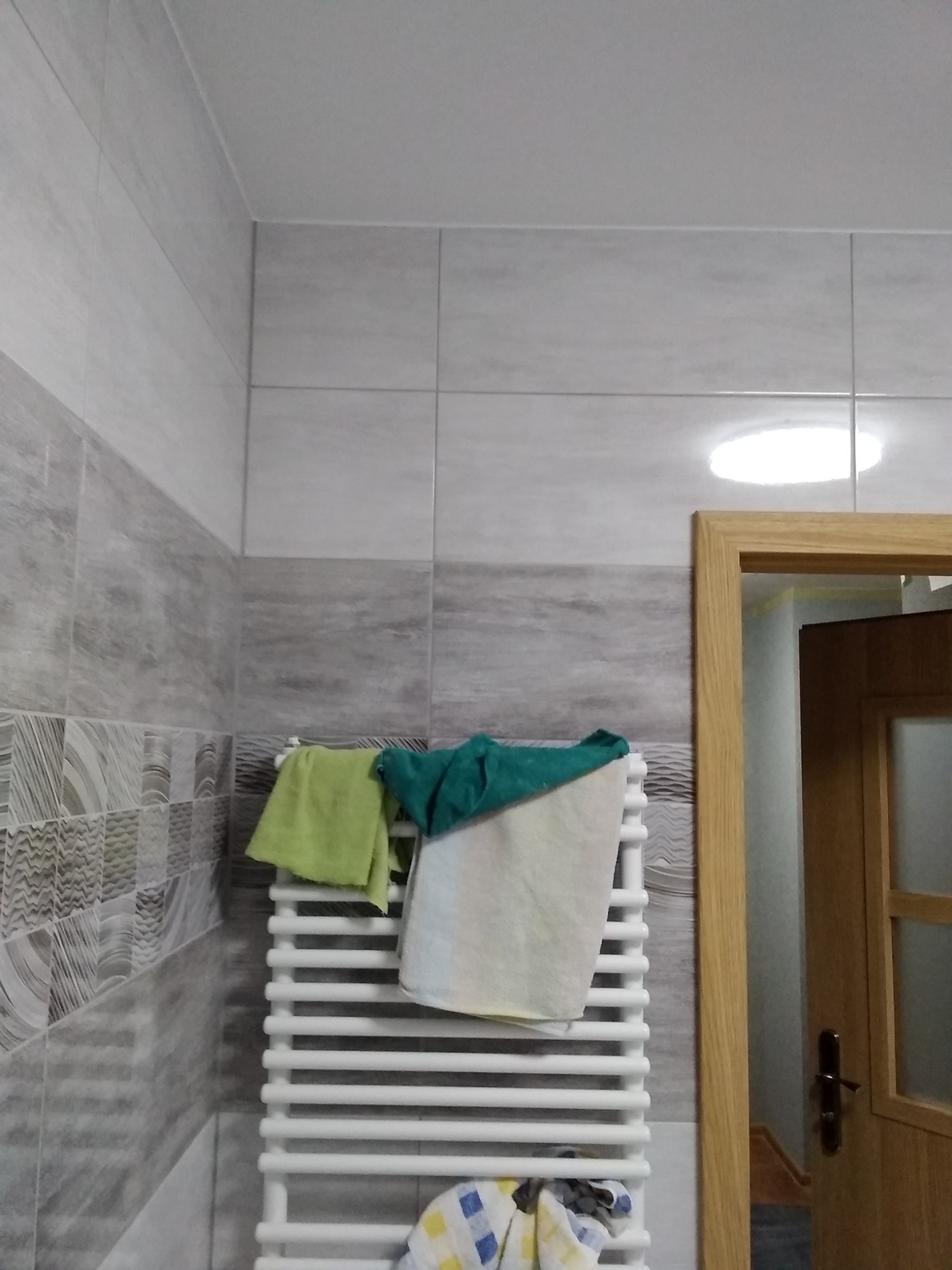 Ściana łazienki z szarymi płytkami w dwóch odcieniach, biały grzejnik drabinkowy z powieszonymi ręcznikami: zielonym, turkusowym i beżowym, fragment drewnianej ościeżnicy drzwiowej.