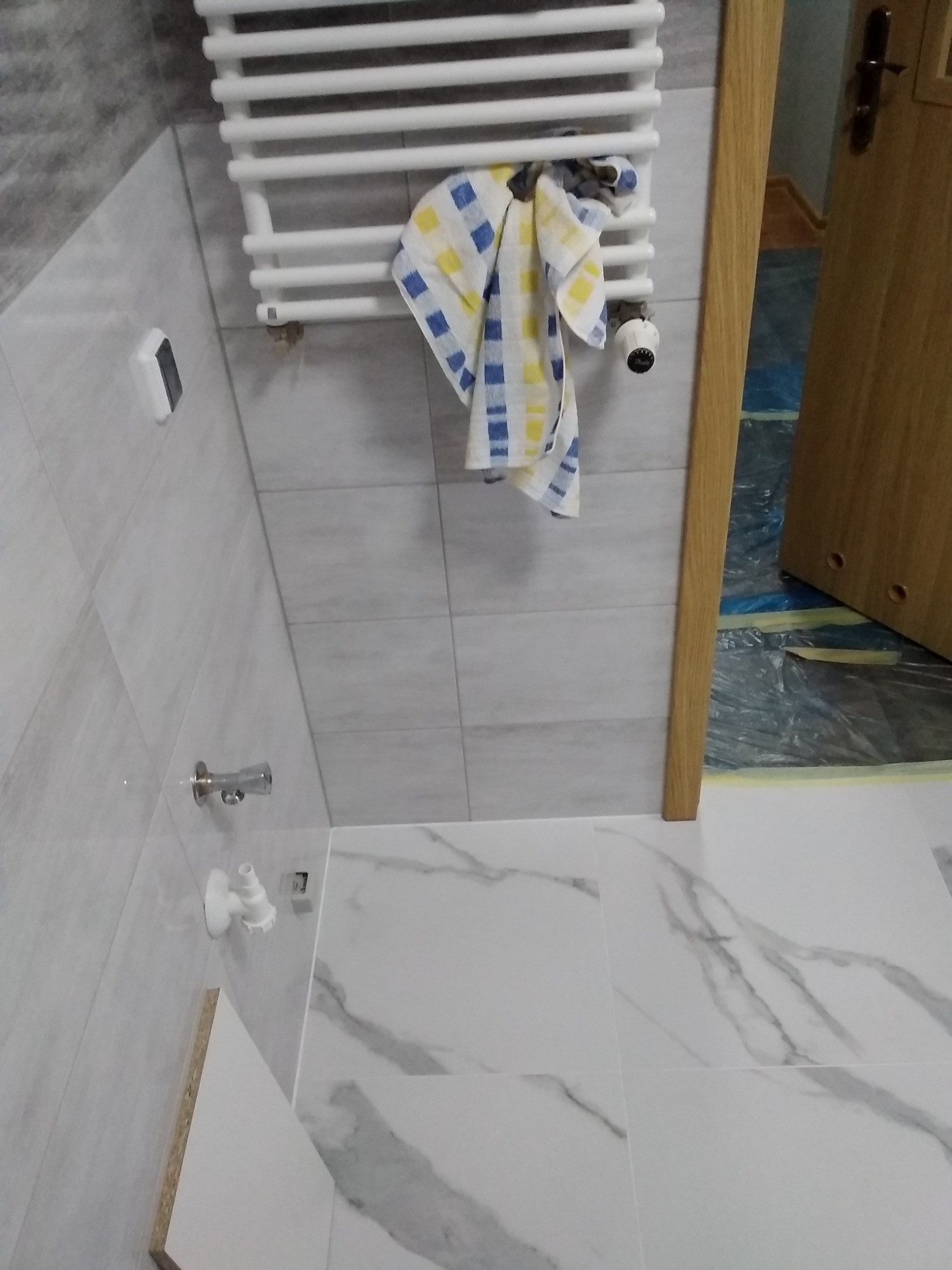 Wykończona łazienka z białymi płytkami imitującymi marmur na podłodze i ścianach, grzejnik drabinkowy z powieszonym ręcznikiem w niebiesko-żółtą kratę, widoczny fragment drzwi i zabezpieczona folią...
