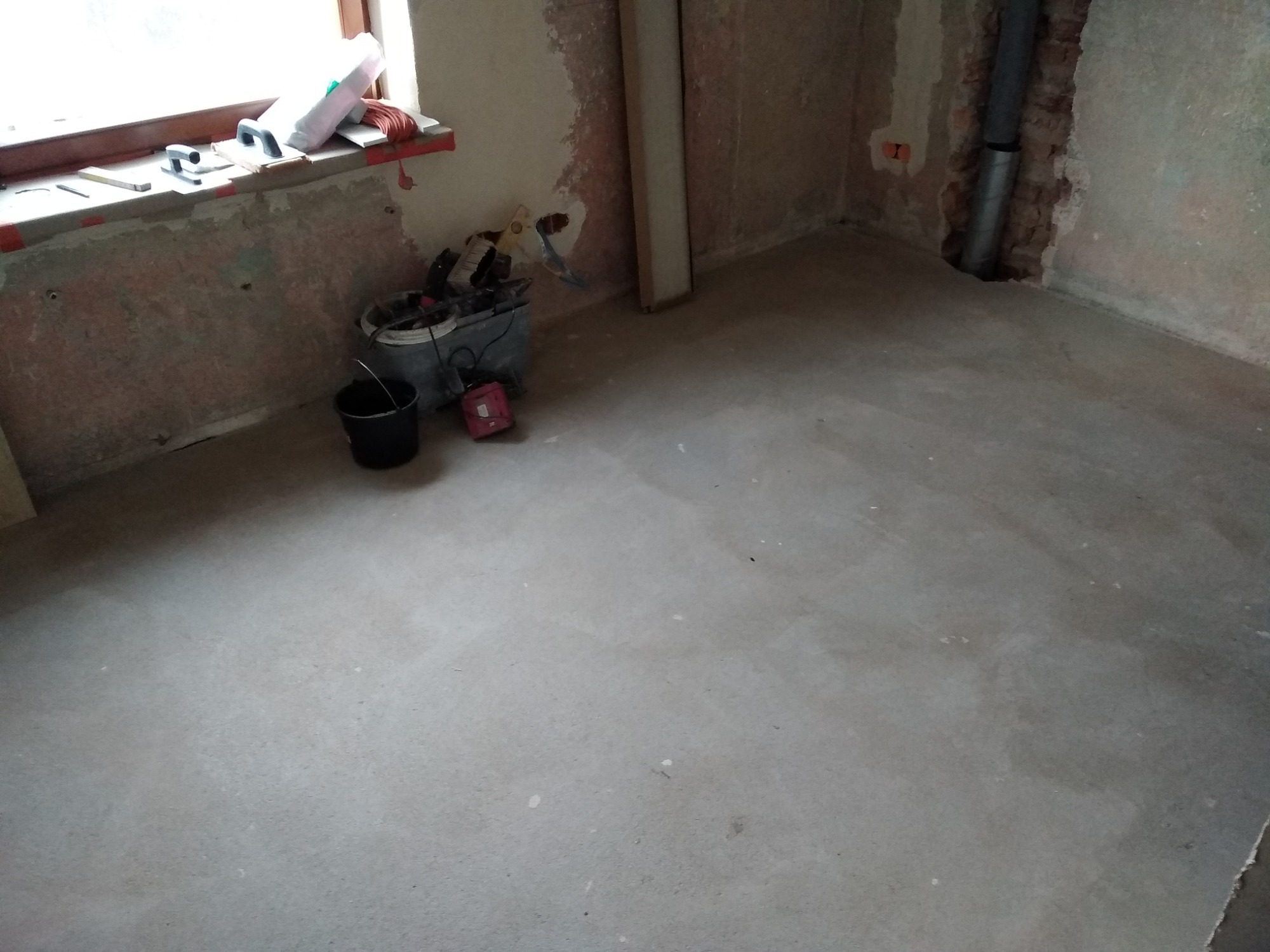 Surowy betonowy wylewka podłogowa w trakcie prac przygotowawczych, widoczne narzędzia i wiadro na parapecie, ściany w stanie surowym, rura kanalizacyjna przy ścianie.