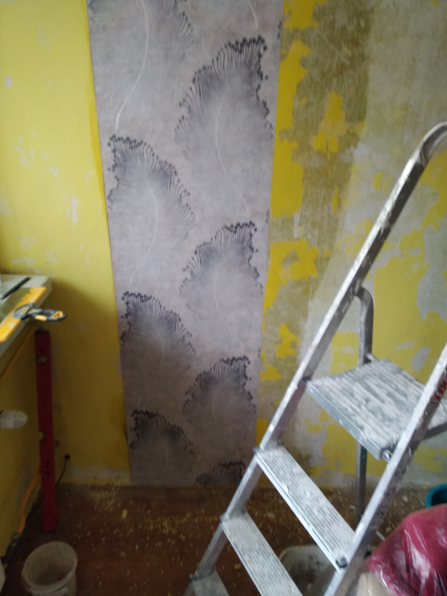Fragment ściany z żółtą farbą i wzorzystą tapetą w trakcie aplikacji, widoczna aluminiowa drabina i narzędzia pomiarowe.
