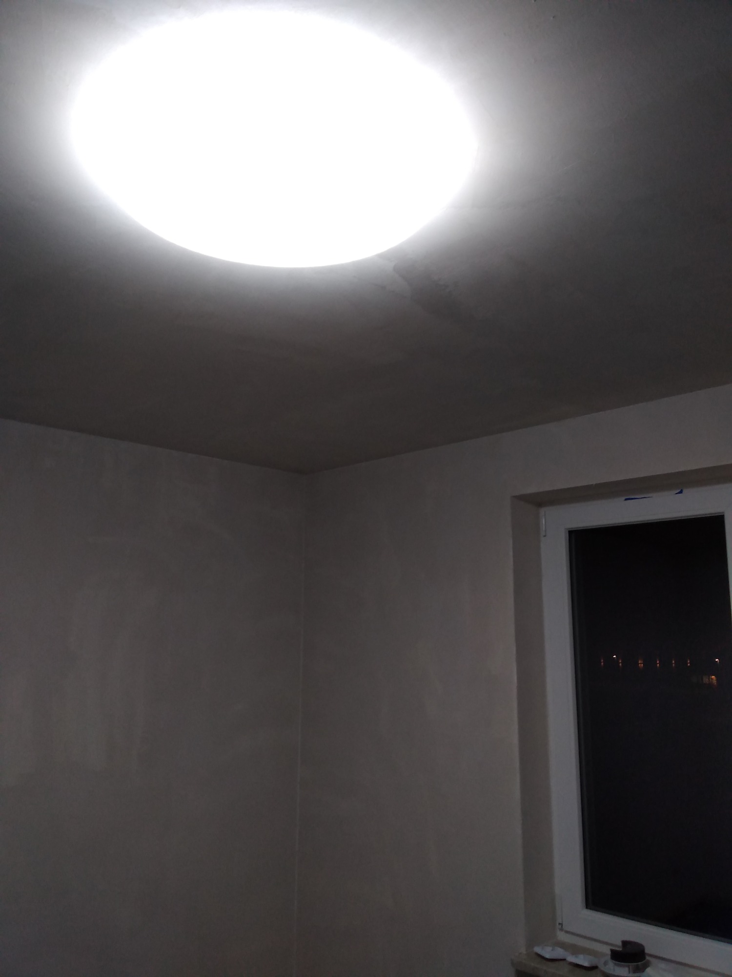 Pokój po szpachlowaniu, widoczna gładka ściana w szarym odcieniu, sufit z okrągłą lampą i okno z widokiem na nocne miasto.