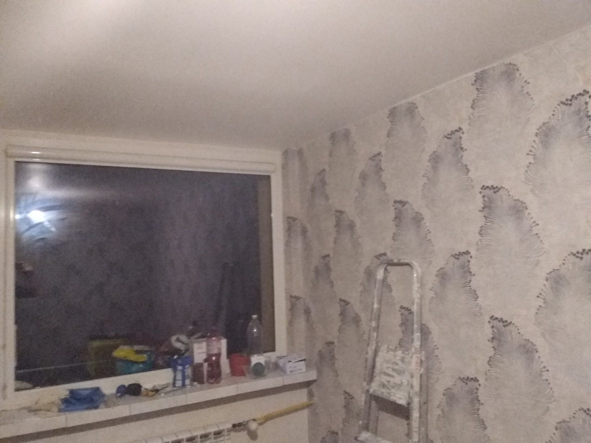 Ściana z tapetą w odcieniach szarości z wzorem przypominającym pióra, fragment okna z parapetem zastawionym różnymi przedmiotami, drabina oparta o ścianę.