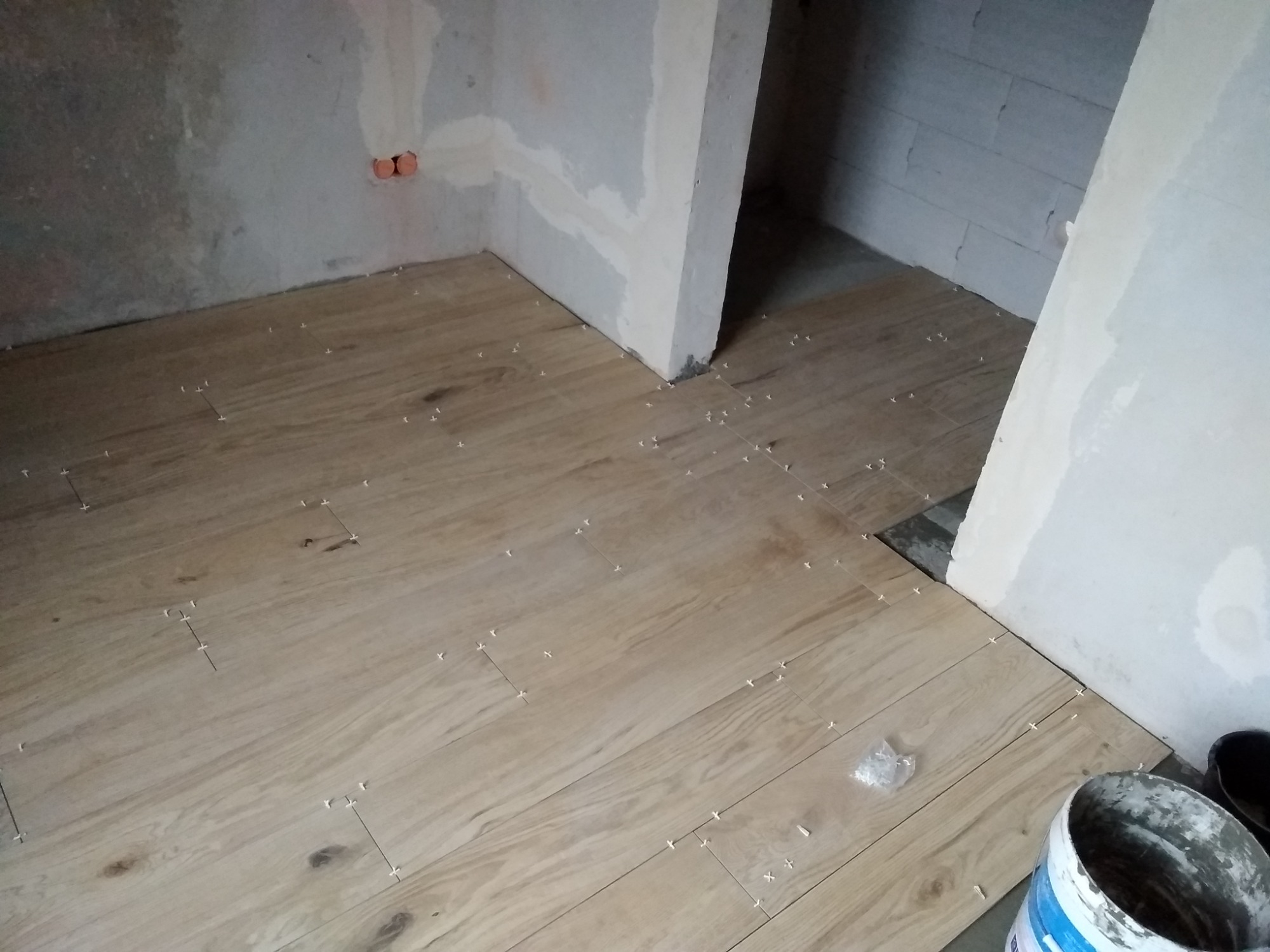 Układanie drewnopodobnych płytek podłogowych w pomieszczeniu w stanie surowym, widoczne krzyżyki dystansowe i wiaderko z klejem.