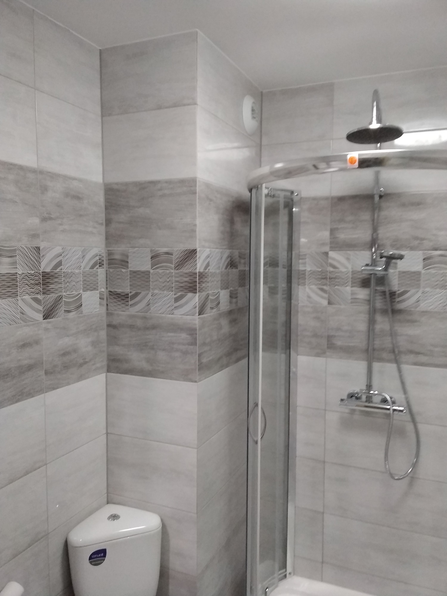 Wykończona łazienka z kabiną prysznicową z przesuwanymi drzwiami, białą toaletą narożną i szarymi płytkami z dekoracyjnym pasem mozaiki.