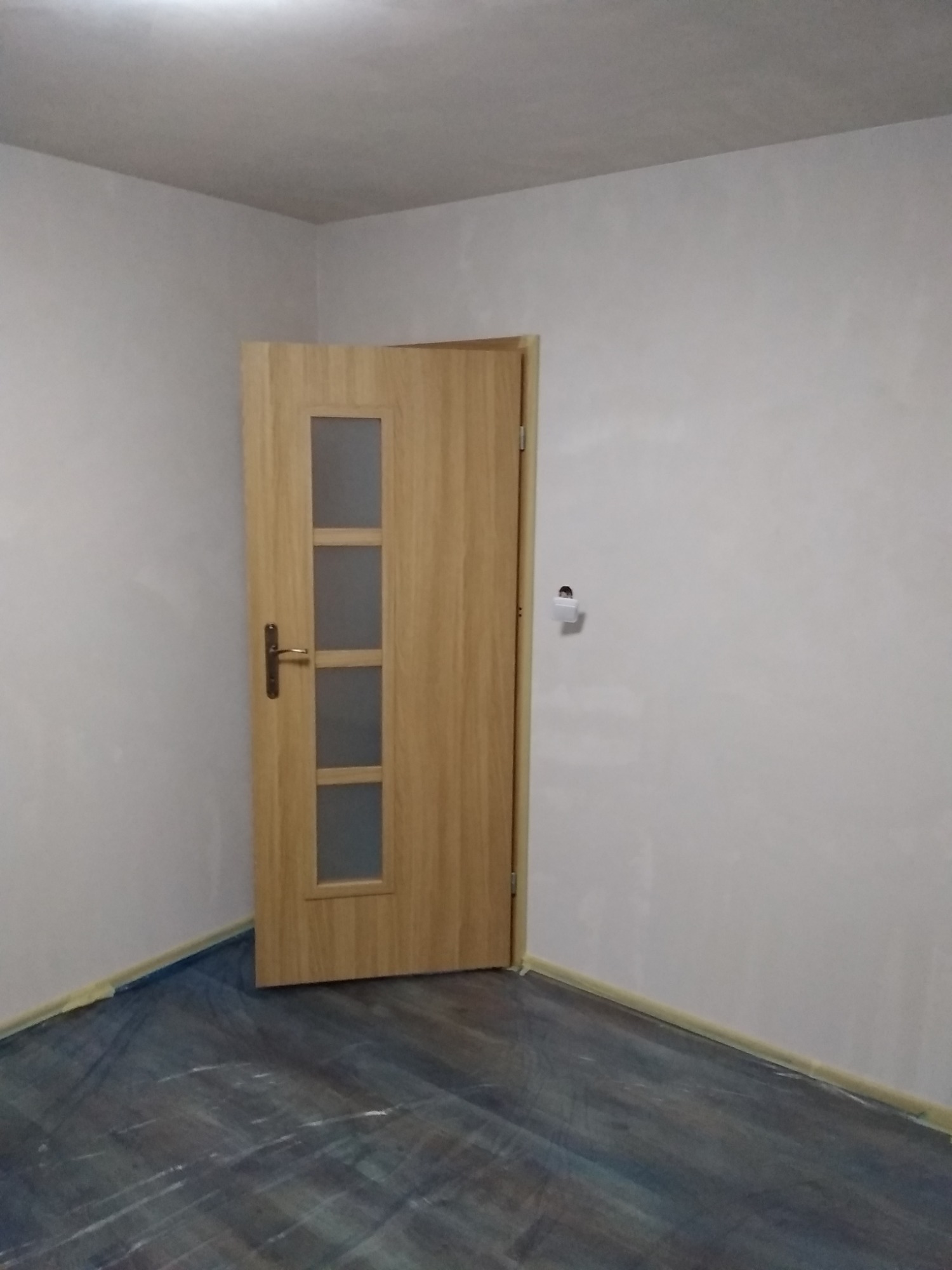 Świeżo pomalowane ściany w pokoju z drewnianą ościeżnicą drzwiową i zabezpieczoną folią podłogą, widoczny fragment listwy przypodłogowej z taśmą malarską.