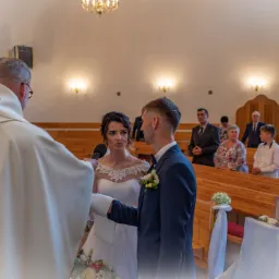 Para młoda podczas ceremonii ślubnej w kościele, ksiądz udziela błogosławieństwa, w tle goście.