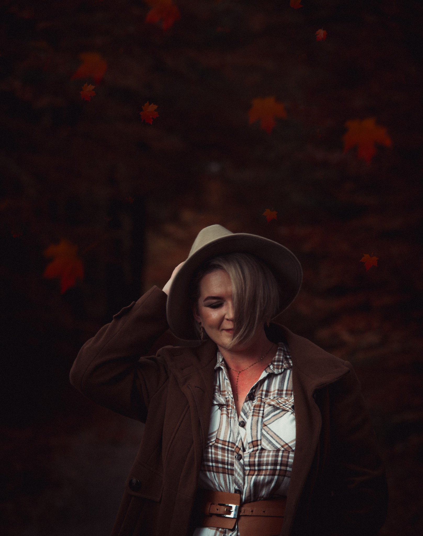 Portret kobiety w brązowym płaszczu i kapeluszu, z opadającymi jesiennymi liśćmi w tle, spoglądającej w dół z lekkim uśmiechem.