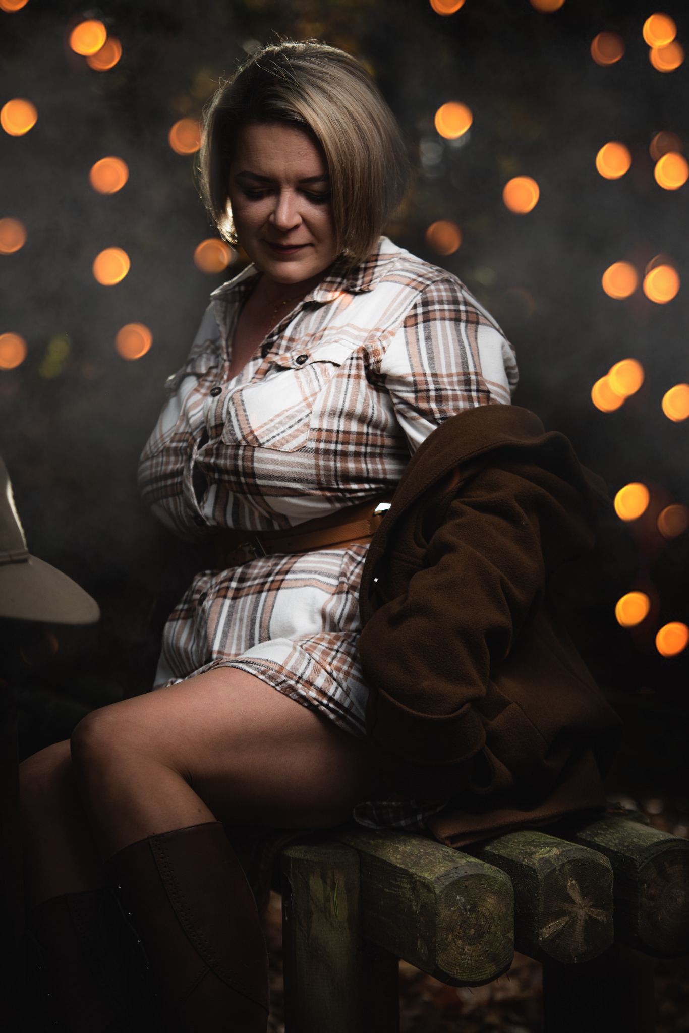 Kobieta siedząca na drewnianej ławce w plenerze, ubrana w koszulową sukienkę w kratę, brązowe kozaki i płaszcz, z rozproszonymi światłami w tle.